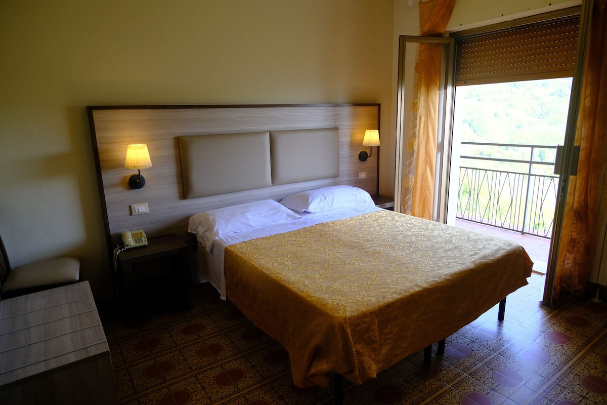A hotel room with a large bed, two bedside lamps, a nightstand with a phone, and a balcony with a view.
