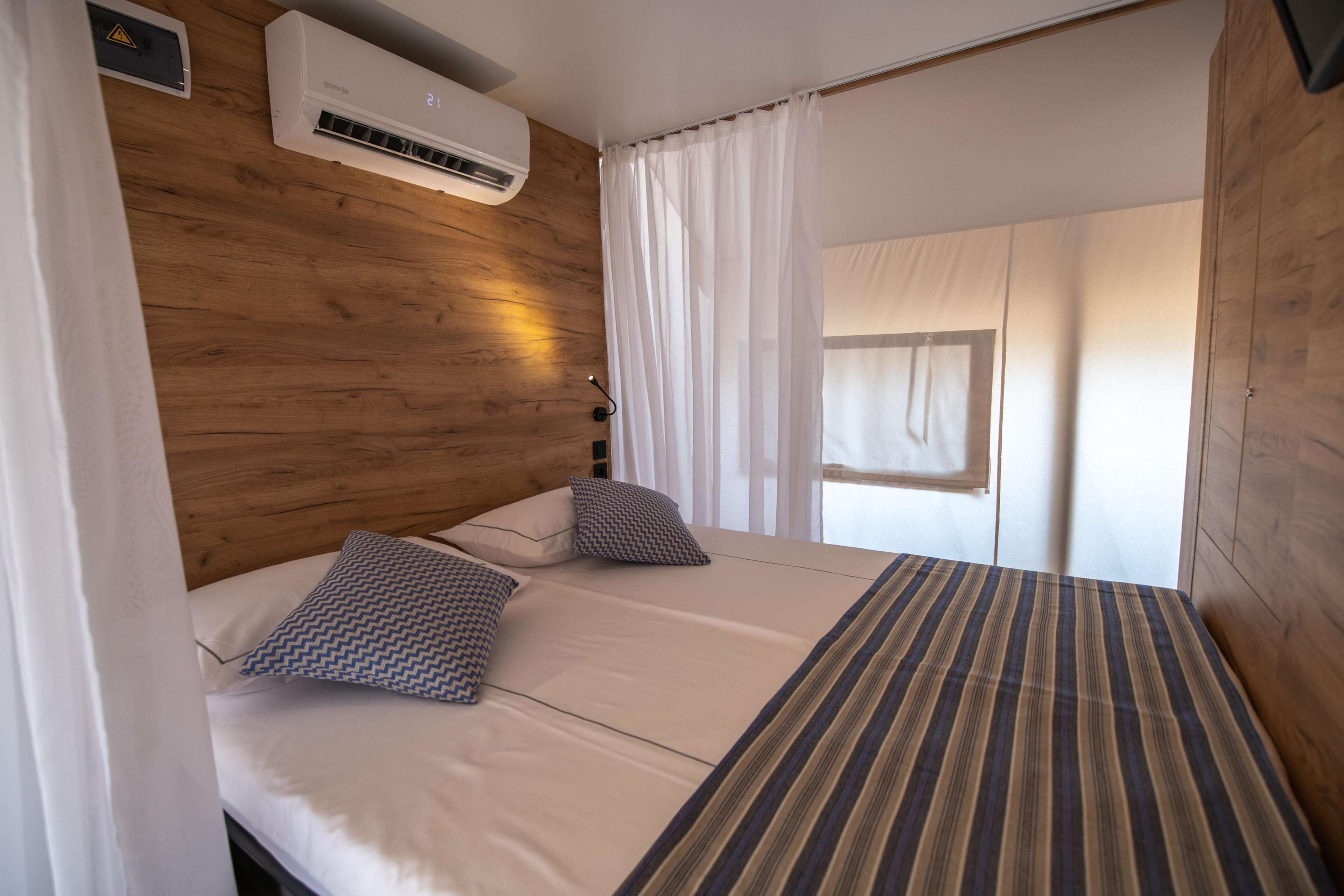 A room with two beds, a wooden headboard, an air conditioning unit, and a window with curtains.