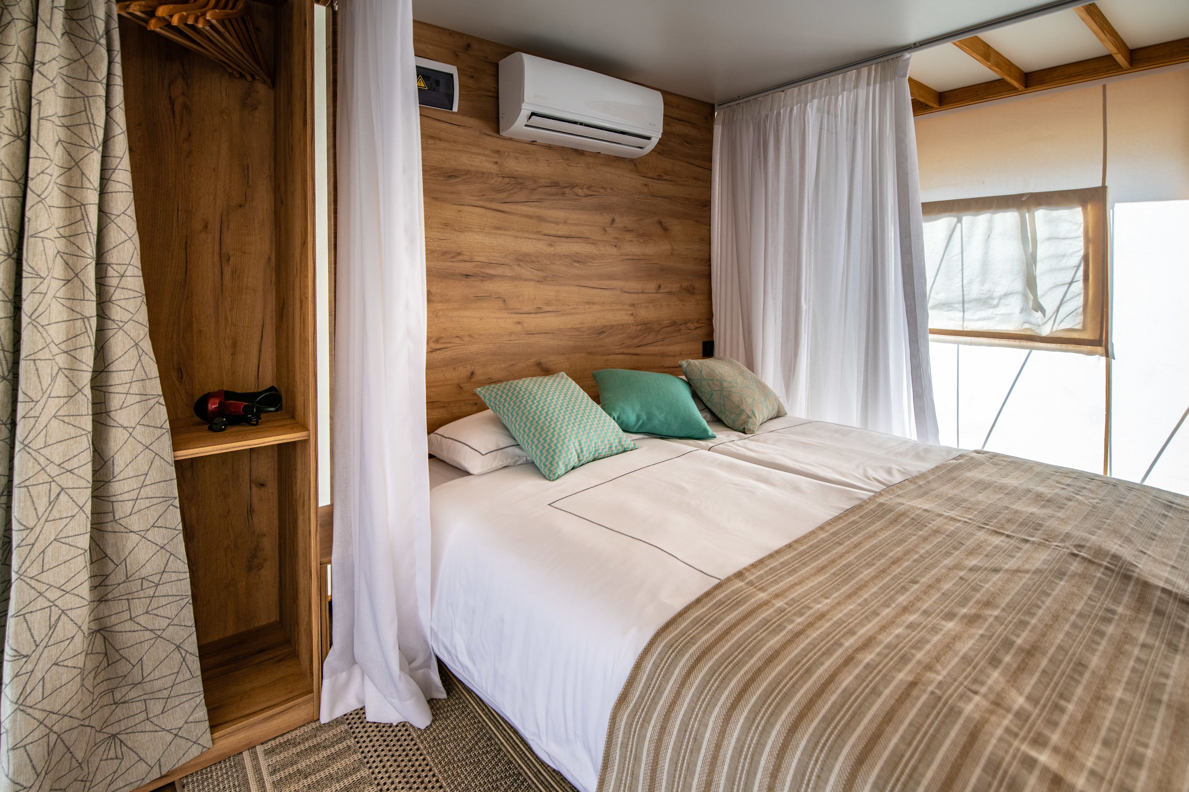 Une chambre à coucher avec une tête de lit en bois, un lit avec une literie blanche et des oreillers à motifs, un appareil de climatisation et une fenêtre avec des rideaux.