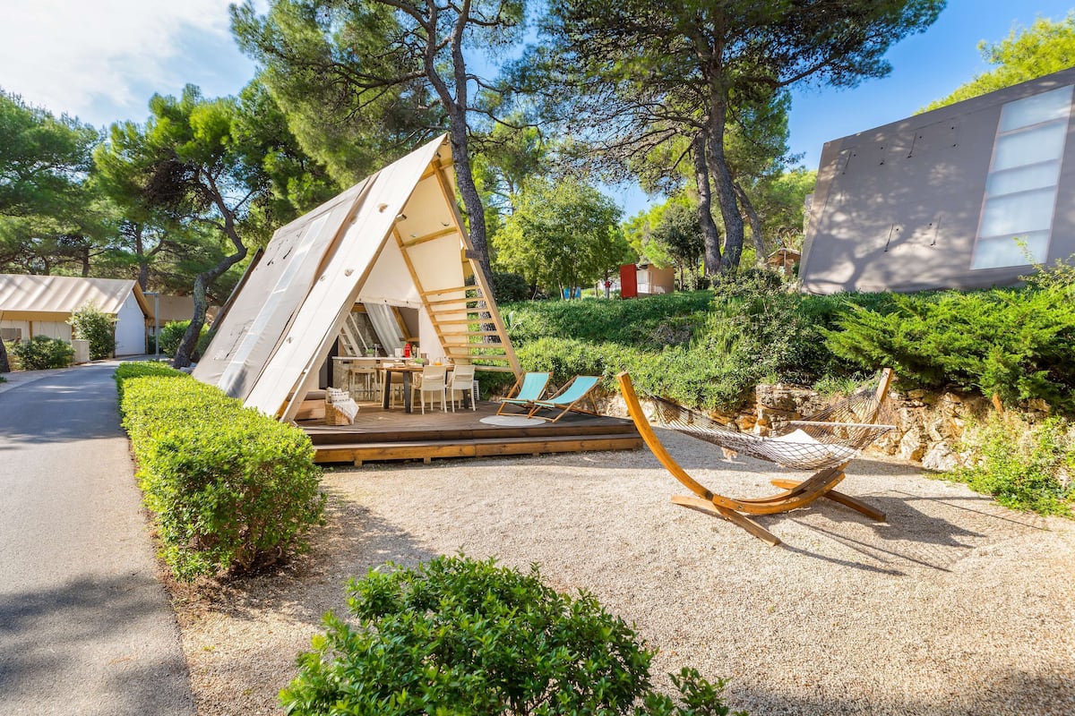 A modern, A-frame wooden structure with a deck and outdoor seating area, surrounded by greenery and a gravel path.