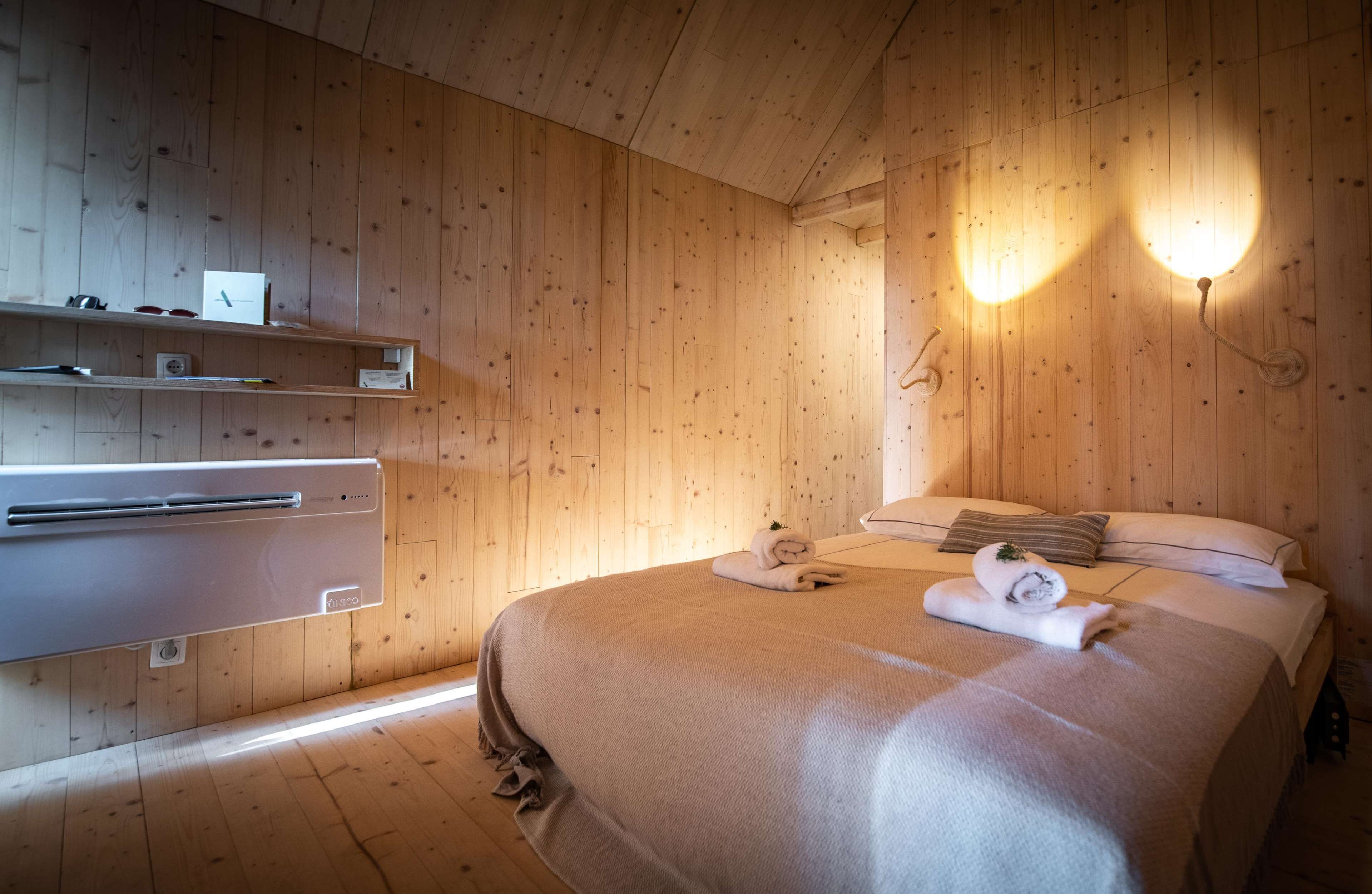 A wooden-paneled room with a bed, two wall-mounted lights, a shelf with items, and a built-in heater.