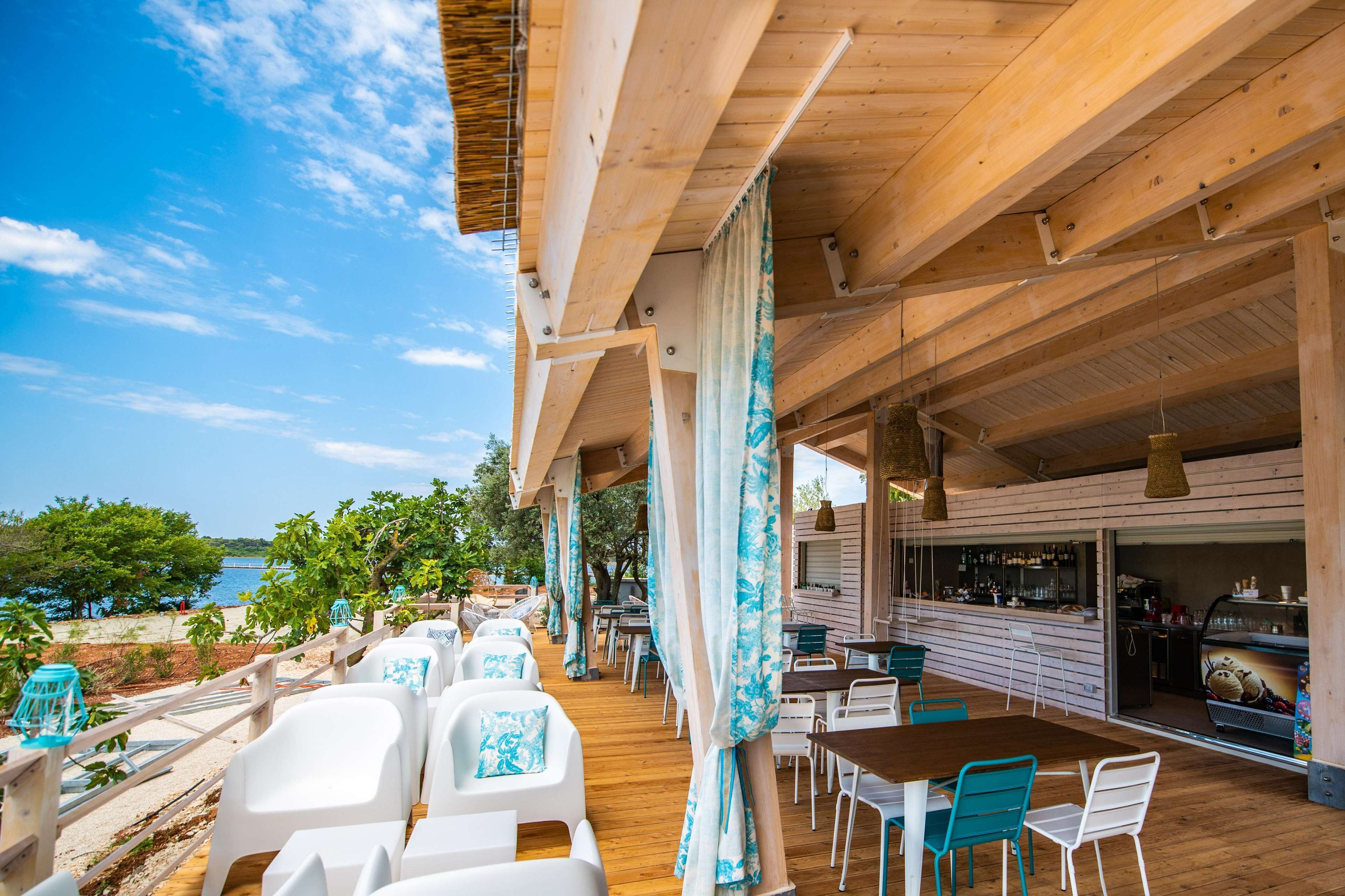 An outdoor dining area with wooden tables and chairs, a bar, and a view of the sea.