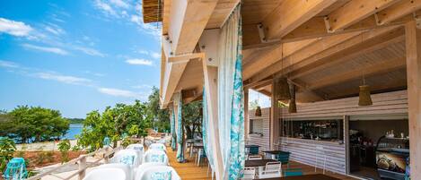 An outdoor dining area with wooden tables and chairs, a bar, and a view of the sea.