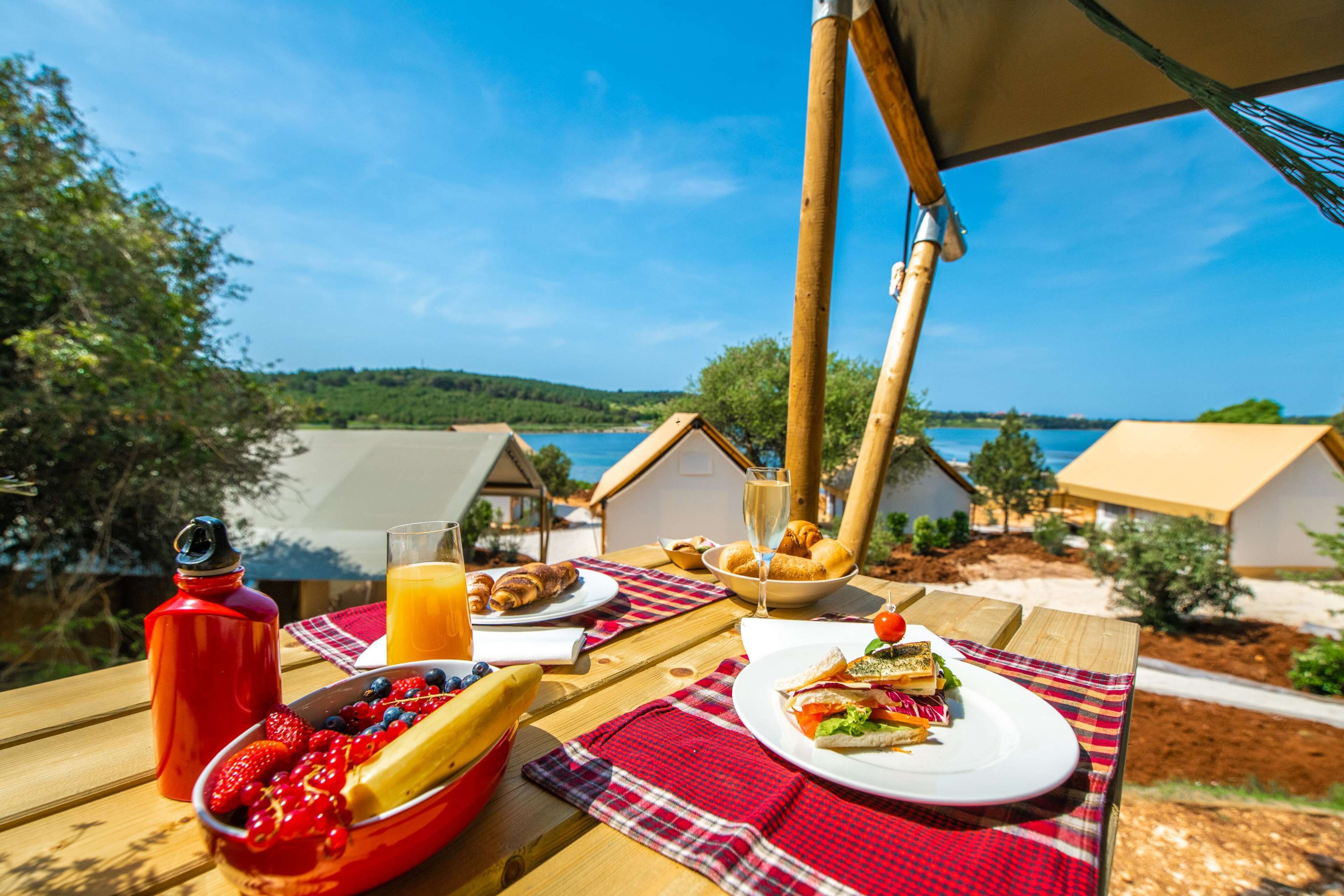 A breakfast table with croissants, a sandwich, and a fruit bowl, overlooking a lake and small houses.