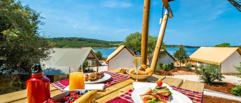A breakfast table with croissants, a sandwich, and a fruit bowl, overlooking a lake and small houses.