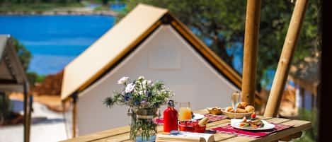 A wooden table set with a book, a glass of juice, a glass of orange juice, a basket of fruit, a plate with food, and a glass of wine.