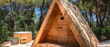 Une structure triangulaire en bois, semblable à une tente, dotée d’une fenêtre circulaire, d’un petit banc et d’un bain à remous, située dans une zone boisée.