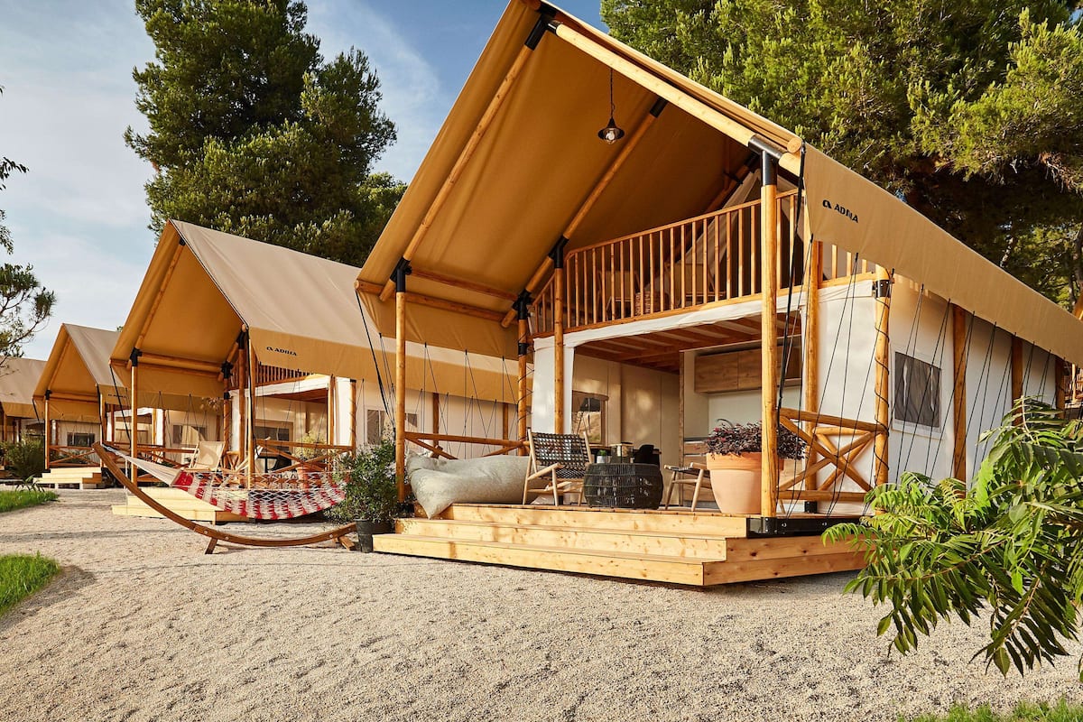 A series of wooden, AADRIA-branded tents with open sides, set on a gravel surface, surrounded by greenery.