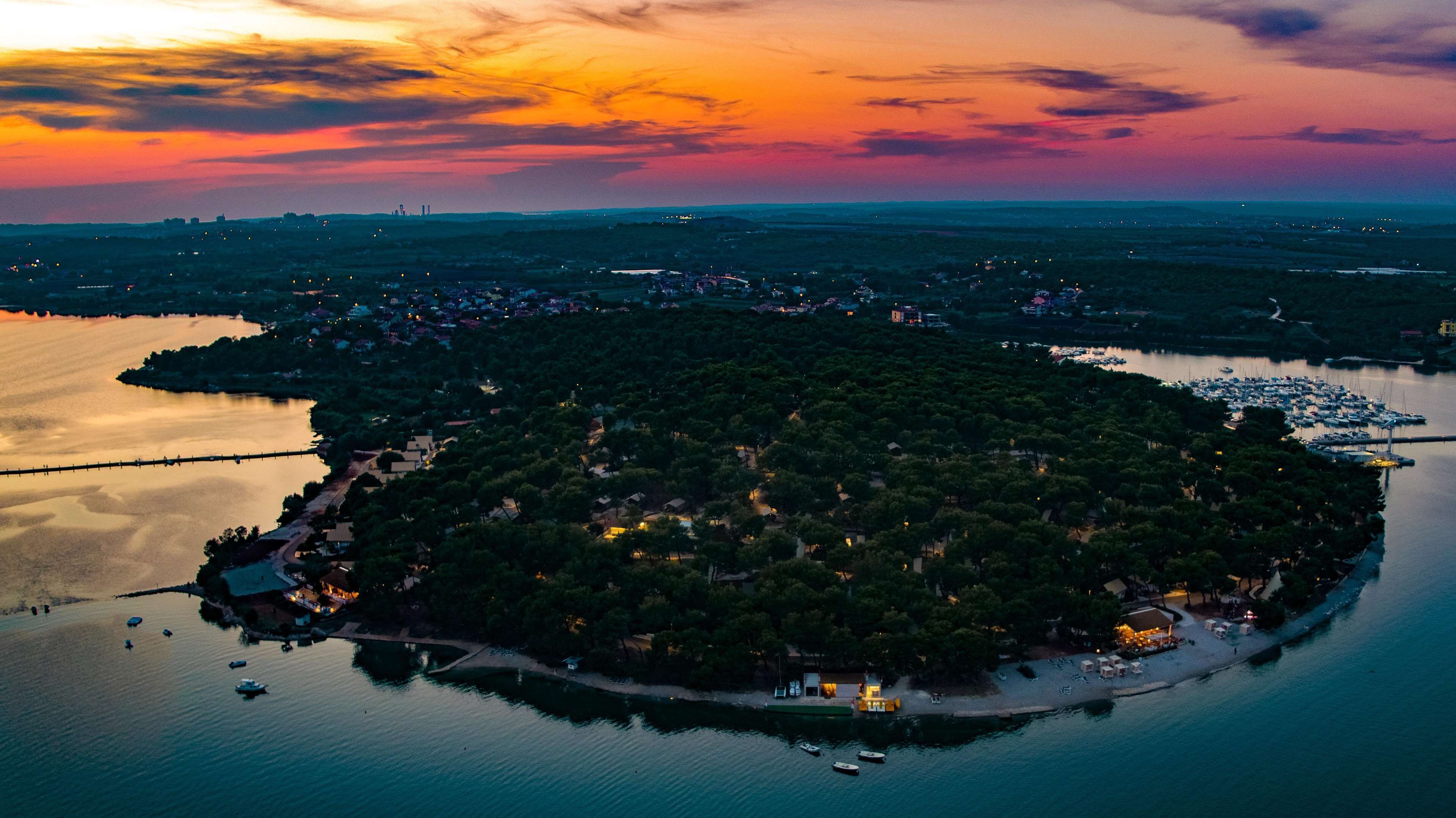 Vue aérienne d’une zone côtière avec une petite île abritant une marina, entourée d’eau et surplombant une ville lointaine.