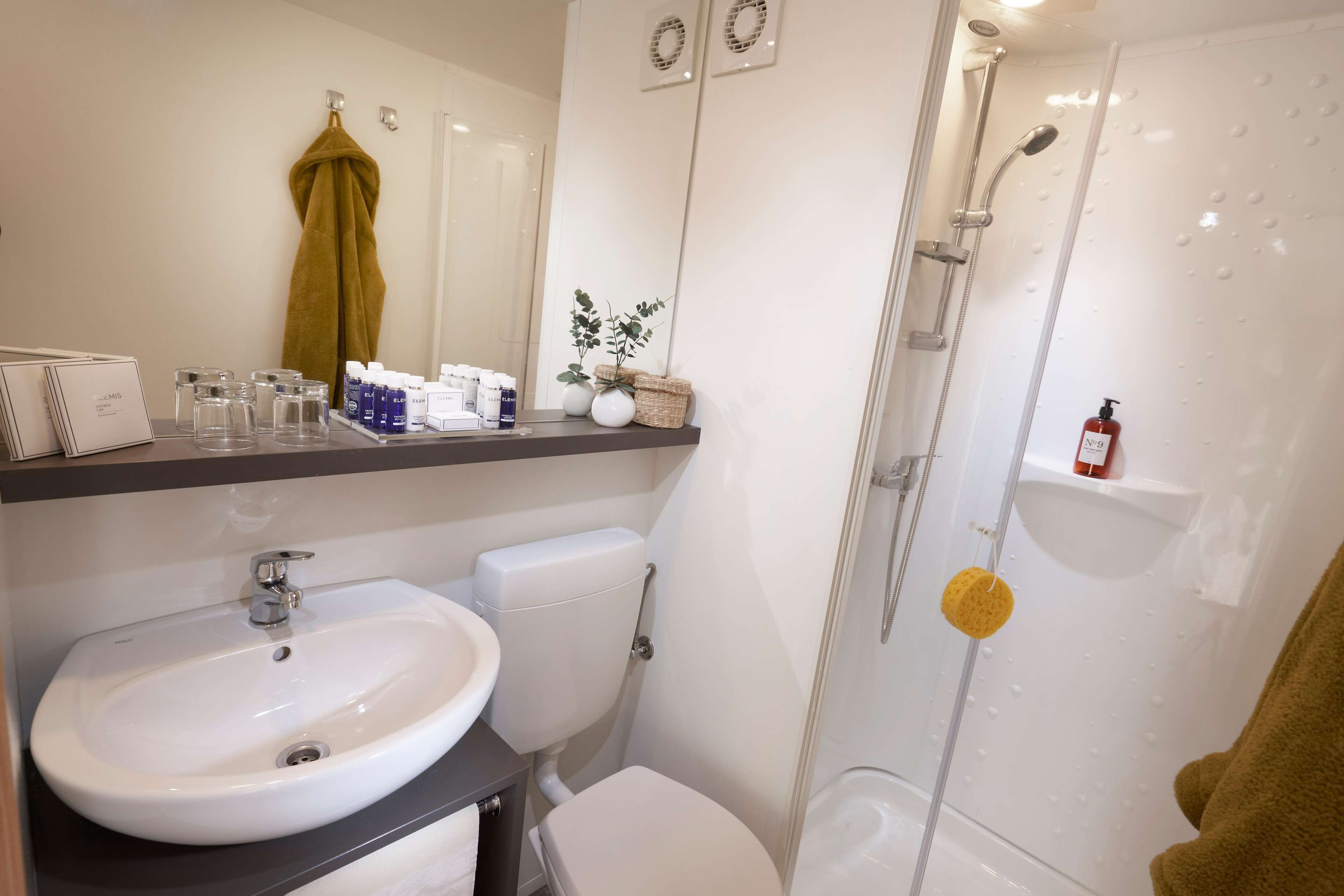 A modern bathroom with a white sink, toilet, and shower. There are toiletries on the counter and a towel hanging on the wall.