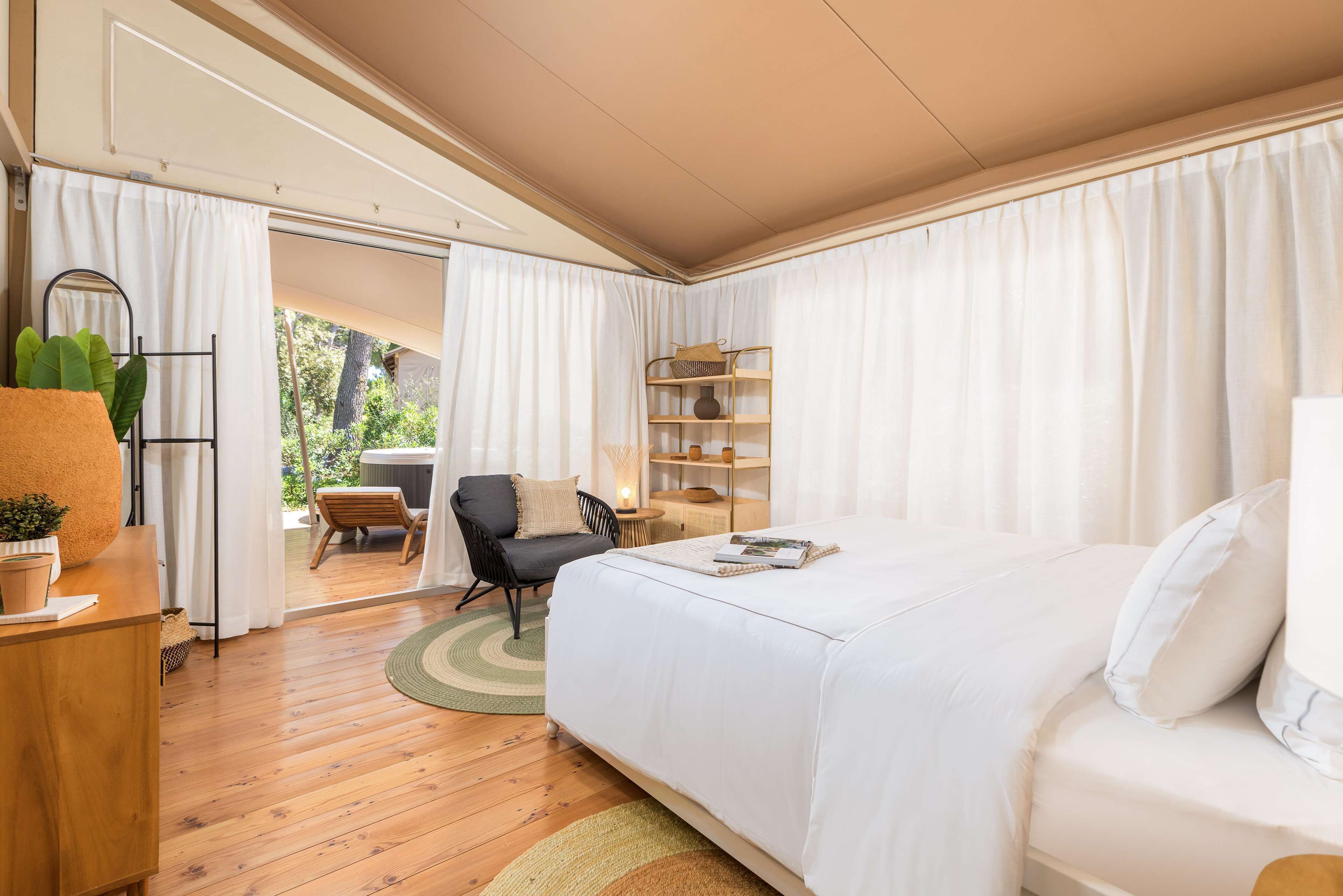 Un dormitorio con una cama grande, un escritorio de madera, una silla y una mesita de noche con lámpara. Se ve una ventana con cortinas y vistas al exterior.