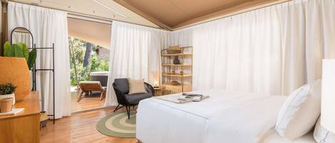 Un dormitorio con una cama grande, un escritorio de madera, una silla y una mesita de noche con lámpara. Se ve una ventana con cortinas y vistas al exterior.