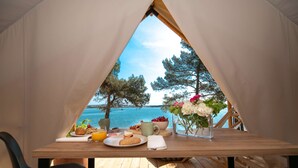 A dining table with breakfast items and a vase of flowers, set against a scenic view of the sea and trees. - Arena One 99 Glamping (Medulin)