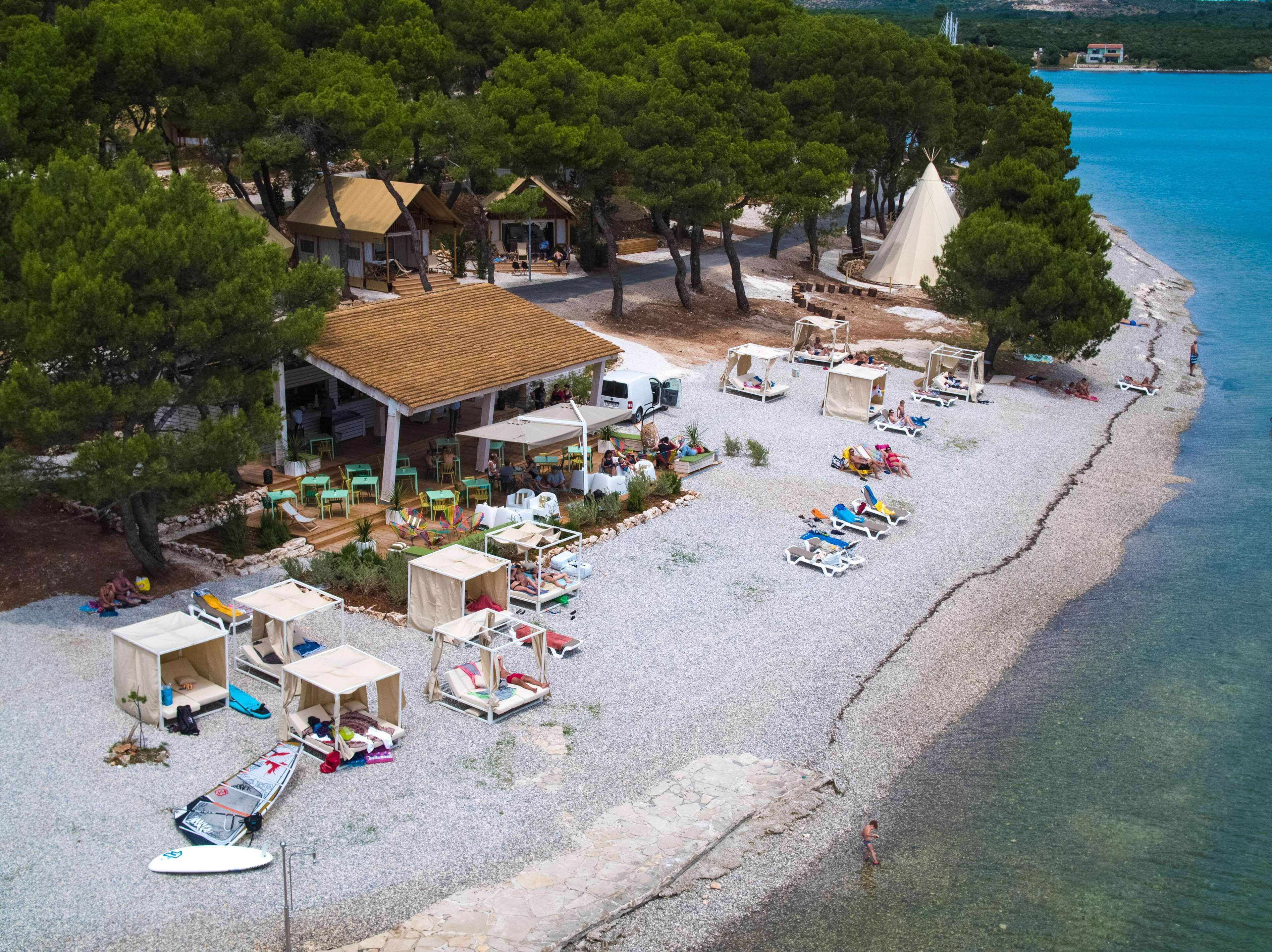 An aerial view of a beachside resort with multiple bungalow-style accommodations, a dining area, and a clear view of the adjacent turquoise sea.