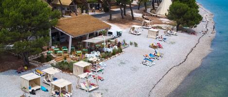 En vy från ovan över ett semesterområde vid stranden med flera bungalovliknande boenden, ett område för måltider och en fri utsikt över det turkosblå havet som ligger intill.