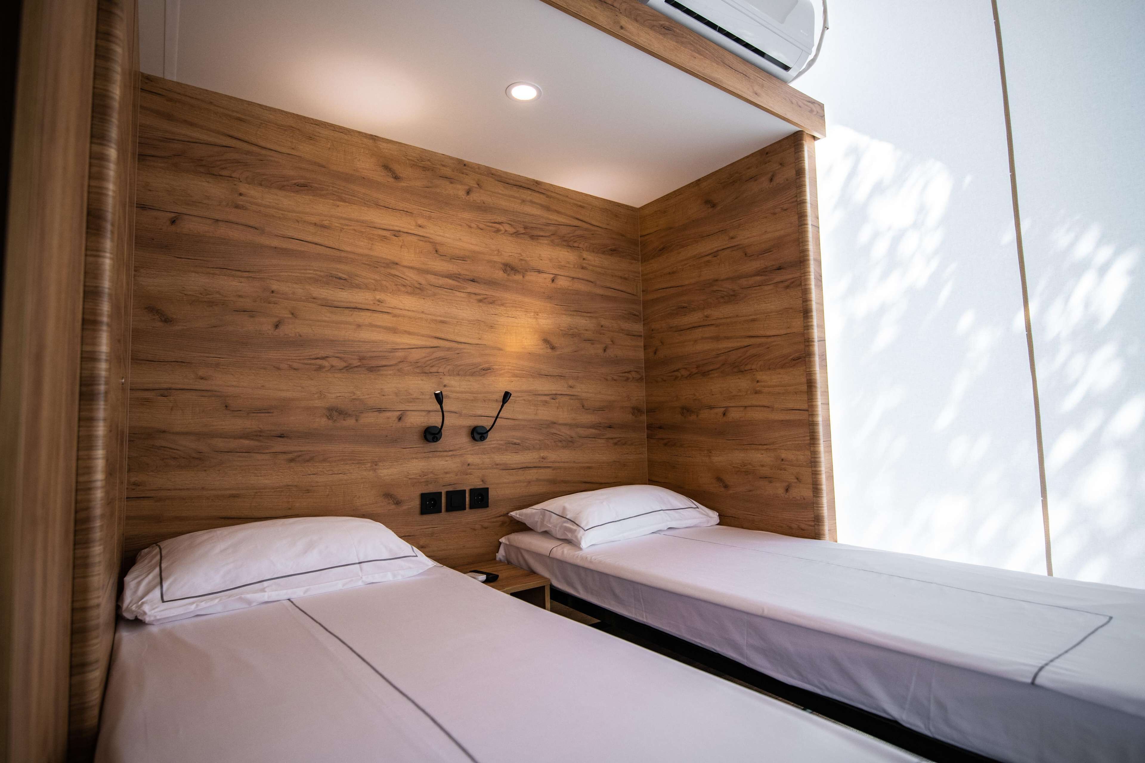 A room with two single beds, wooden panel walls, a ceiling light, and a window.