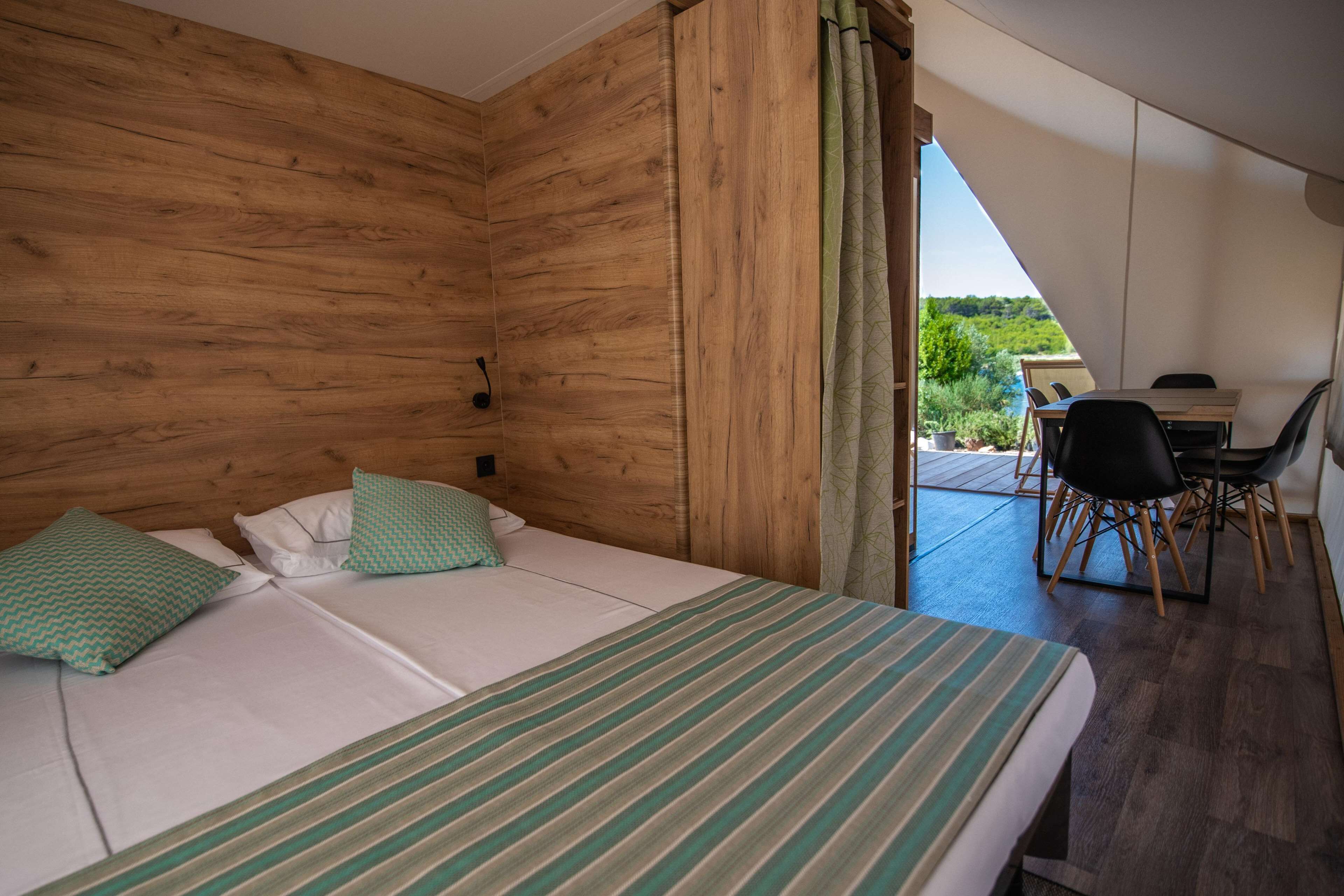 A bedroom with a bed, two pillows, a wooden wall, a desk with a chair, and a view of greenery.