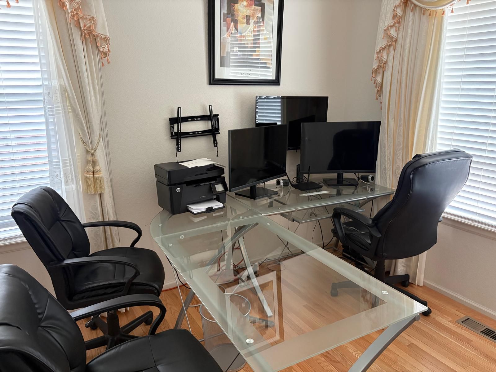 Um espaço de escritório moderno com uma mesa de vidro, cadeiras de escritório pretas, dois monitores, uma impressora e um quadro na parede.