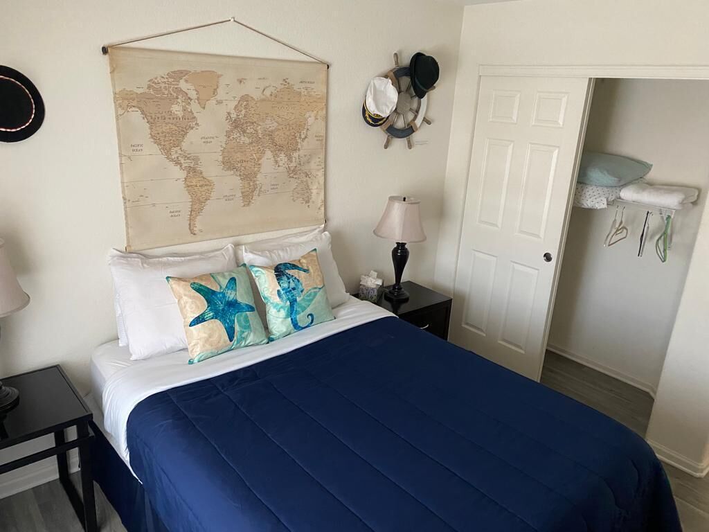A neatly made bed with a blue bedspread and starfish pillows, a world map on the wall, a ship's wheel, and a door leading to a bathroom with towels and toiletries.