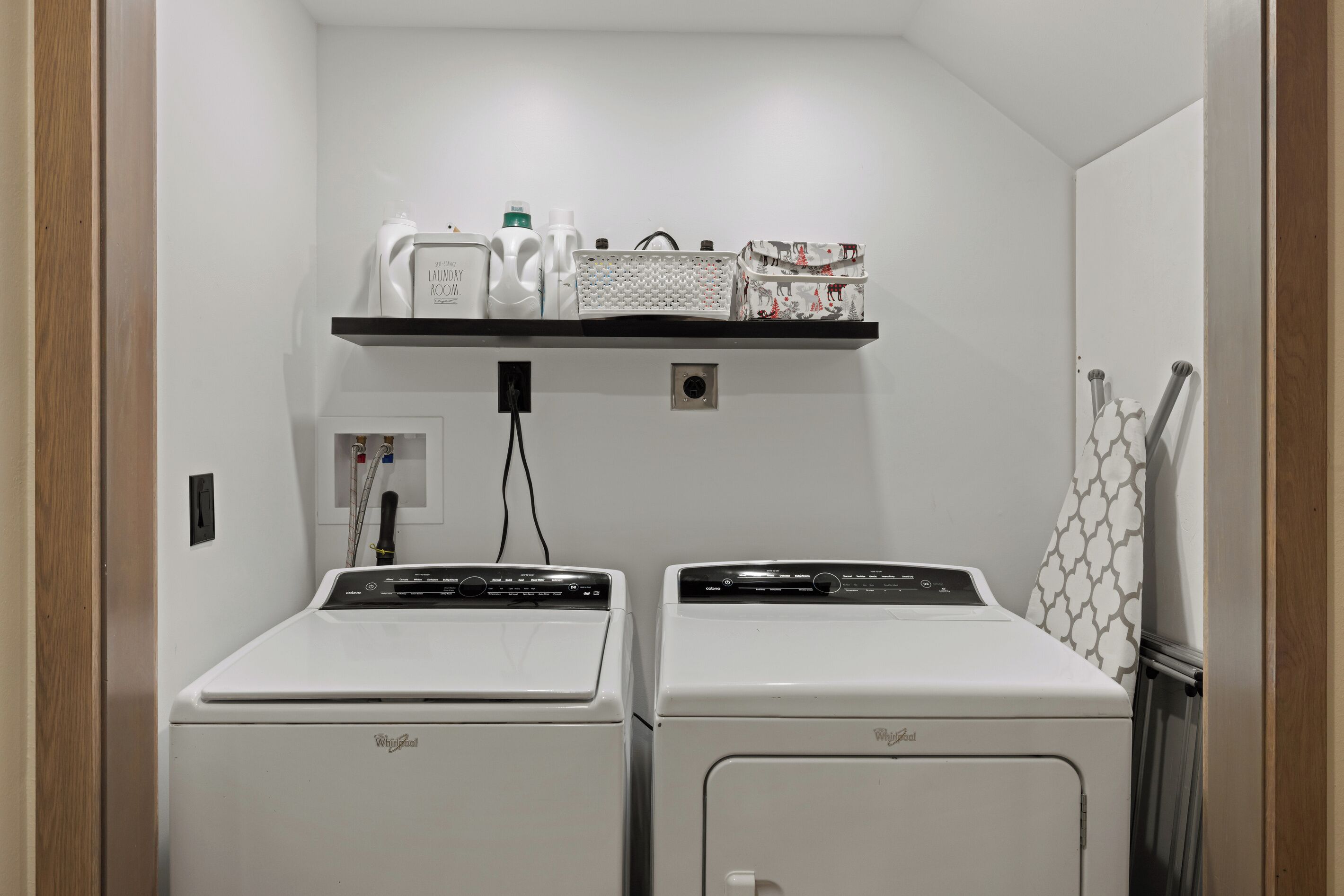 A laundry room with two white Whirlpool washers and dryers, a shelf with laundry supplies, and a patterned towel rack.