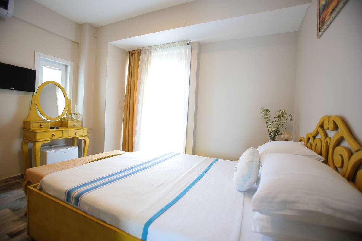 A hotel room with a bed, a wooden headboard, a bedside table with a mirror, and a small window with curtains.
