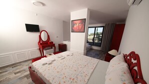 Habitación de hotel con cama, cómoda roja, espejo, televisor de pantalla plana y ventana con vista al exterior.