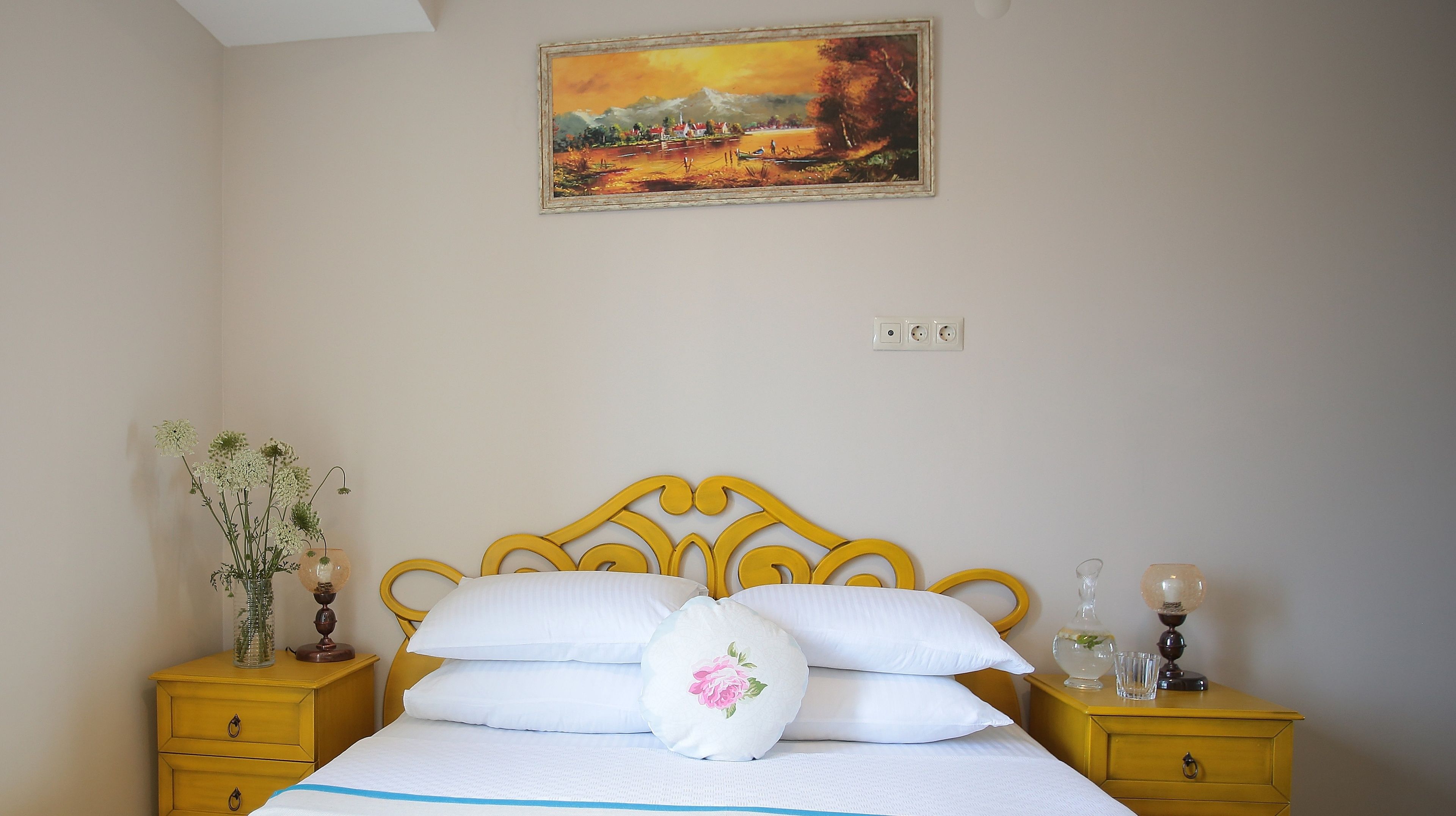 A bedroom with a bed, two nightstands, a vase with flowers, a painting on the wall, and a window.