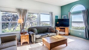 A living room with a sofa, coffee table, TV, and large windows with curtains. - Ocean Front Home With Hot Tub On Miles Of Sandy Beach! (Yachats)