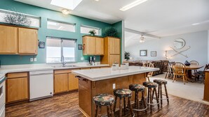 A modern kitchen with wooden cabinets, a central island with stools, and a dining area with a round table and chairs. - Ocean Front Home With Hot Tub On Miles Of Sandy Beach! (Yachats)