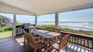 A covered outdoor deck with a wooden table and chairs, overlooking a beach. - Ocean Front Home With Hot Tub On Miles Of Sandy Beach! (Yachats)