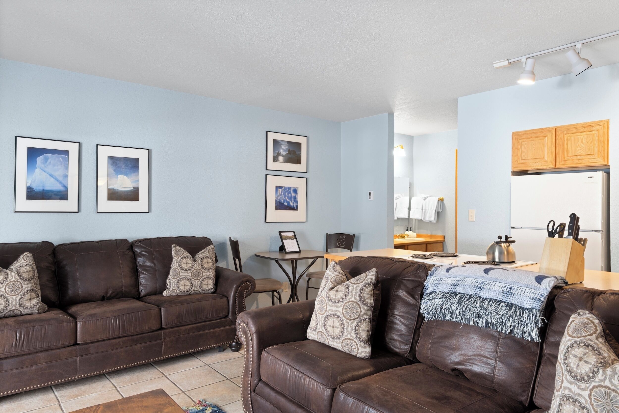 A living room with two brown leather sofas, a coffee table, and a dining area with a table and chairs. There are framed pictures on the wall and a kitchen area with wooden cabinets and a refrigerator.