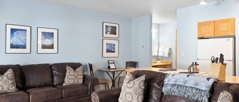 A living room with two brown leather sofas, a coffee table, and a dining area with a table and chairs. There are framed pictures on the wall and a kitchen area with wooden cabinets and a refrigerator.