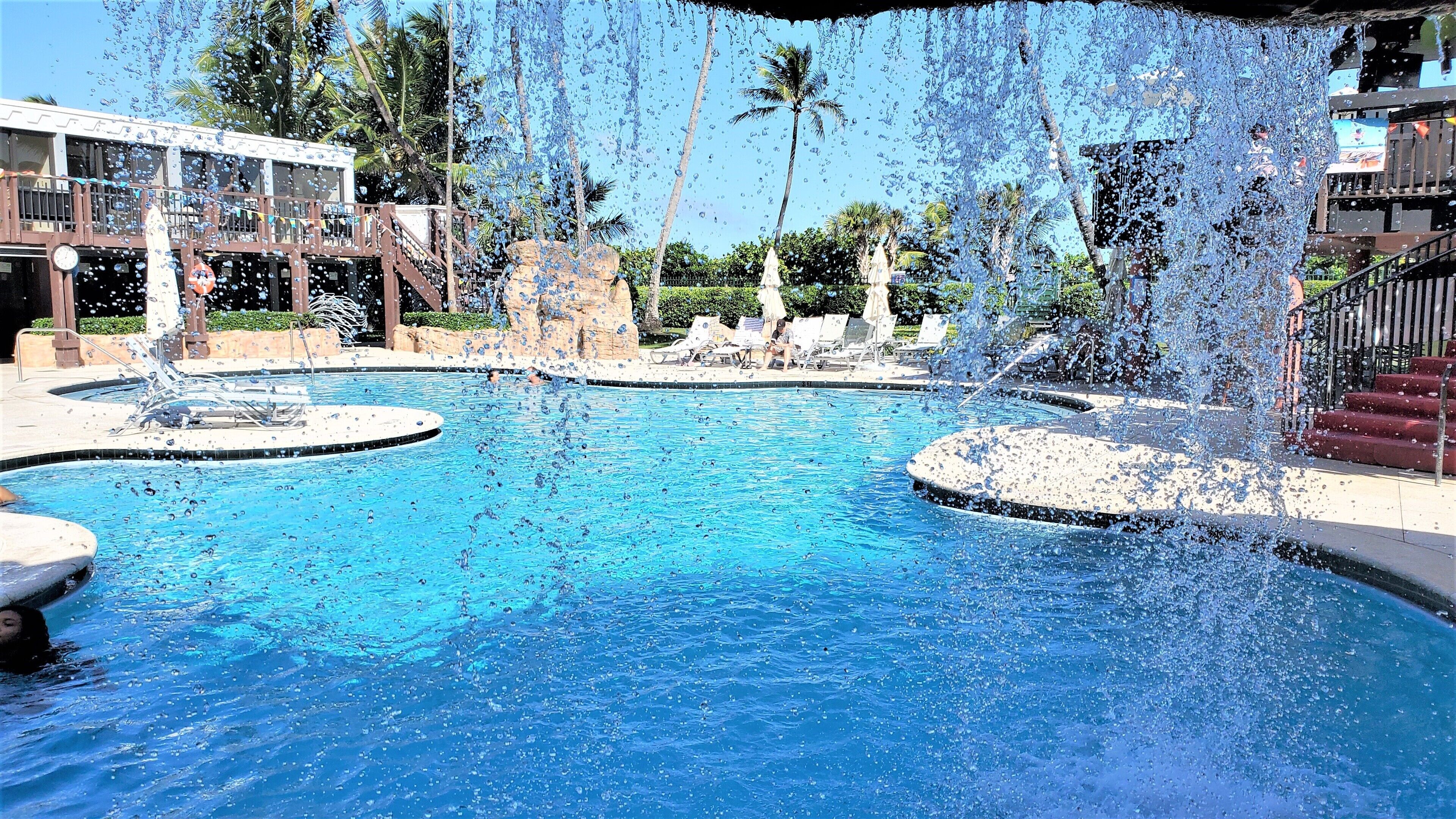 Una piscina con chorros de agua, sillas de descanso y palmeras al fondo.