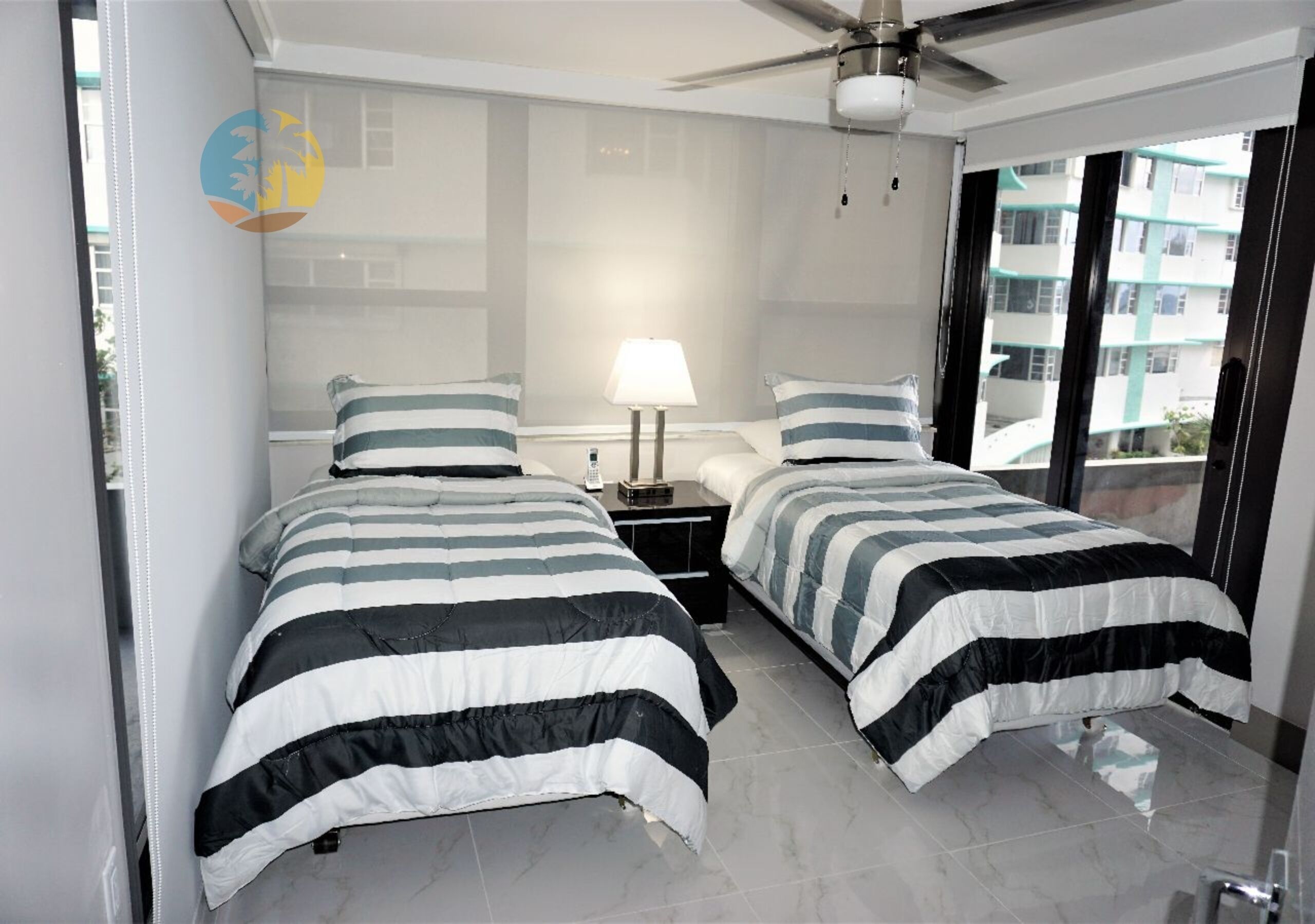 Une chambre avec deux lits, un ventilateur de plafond, une table de chevet avec une lampe et une vue sur l’extérieur par une fenêtre.