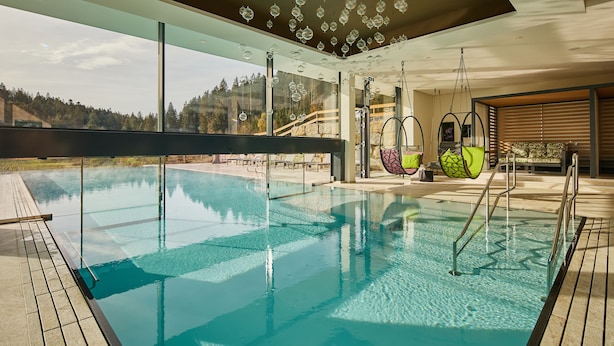 Una piscina interior moderna con paredes de vidrio transparente, un suelo de madera y vistas a una ladera boscosa.