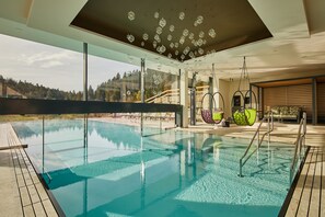 A modern indoor swimming pool with clear glass walls, a wooden deck, and a view of a forested hillside. - Hotel Edita (Scheidegg)