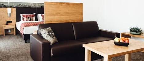 Una habitación de hotel moderna con un sofá marrón oscuro, un cabecero de madera, una pequeña mesa con un frutero y una cama con una colcha de cuadros rojos y blancos.