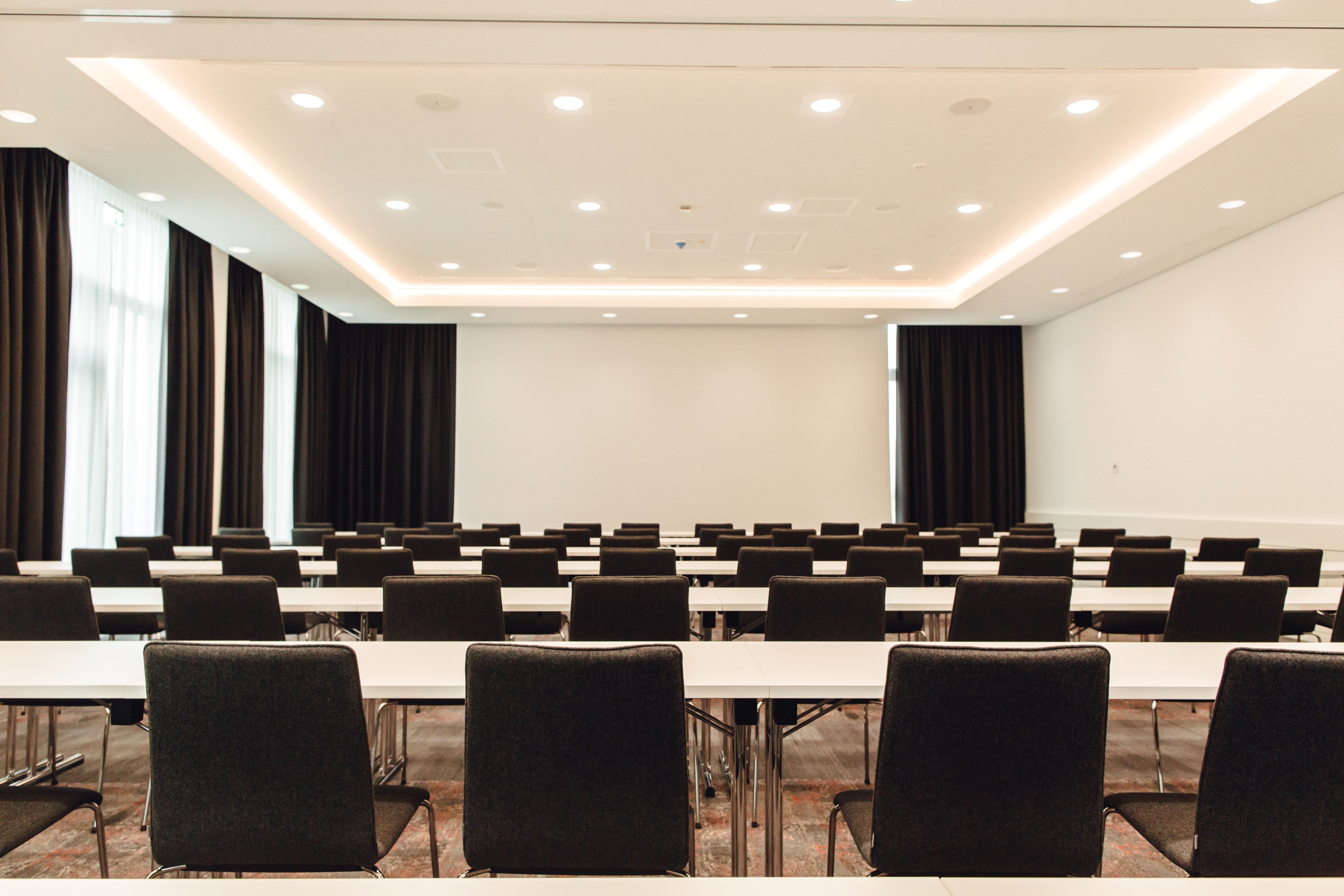 Une salle de conférence avec des rangées de tables et de chaises, de grandes fenêtres avec des rideaux et un tableau blanc au mur.