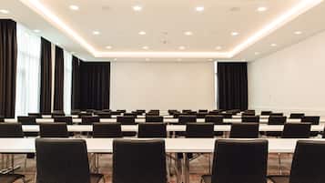 Une salle de conférence avec des rangées de tables et de chaises, de grandes fenêtres avec des rideaux et un tableau blanc au mur.