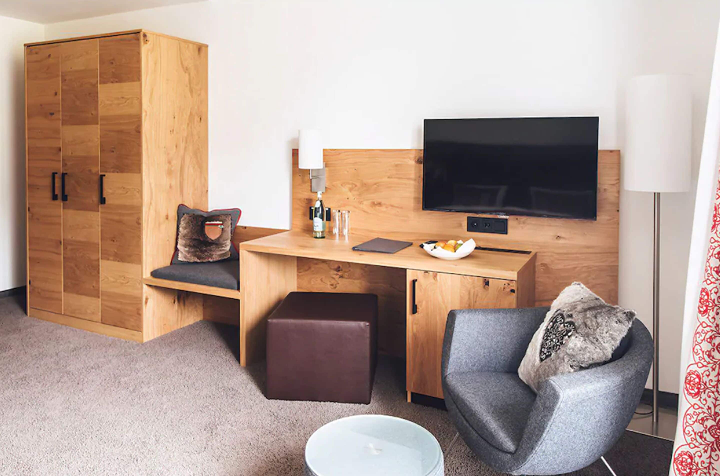 A compact hotel room with a wooden wardrobe, a desk with a flat-screen TV, a grey armchair, and a small ottoman.