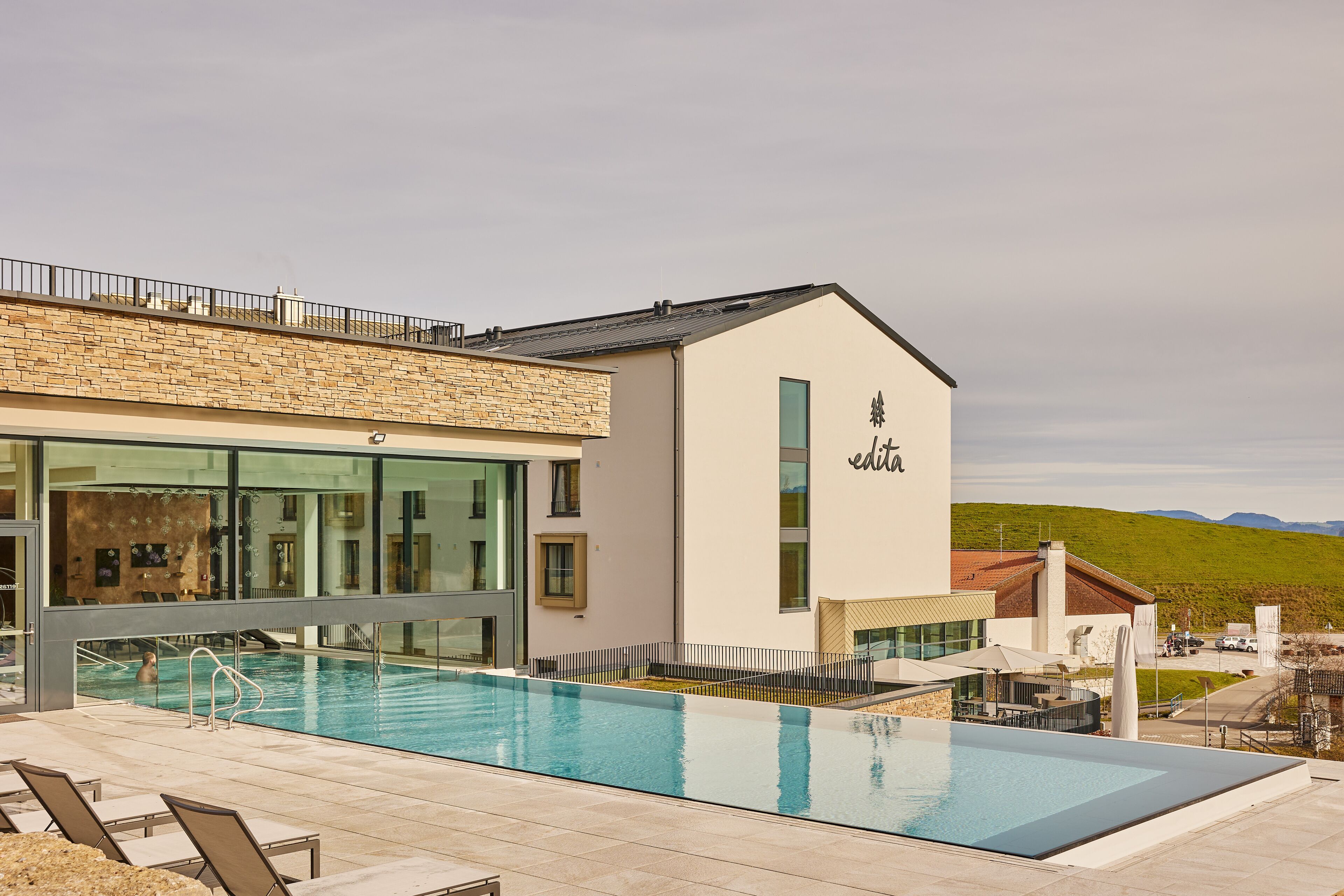 A modern building with a swimming pool, outdoor seating, and a view of the countryside.