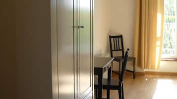A room with a large white wardrobe, a small table with a chair, and a window with curtains.