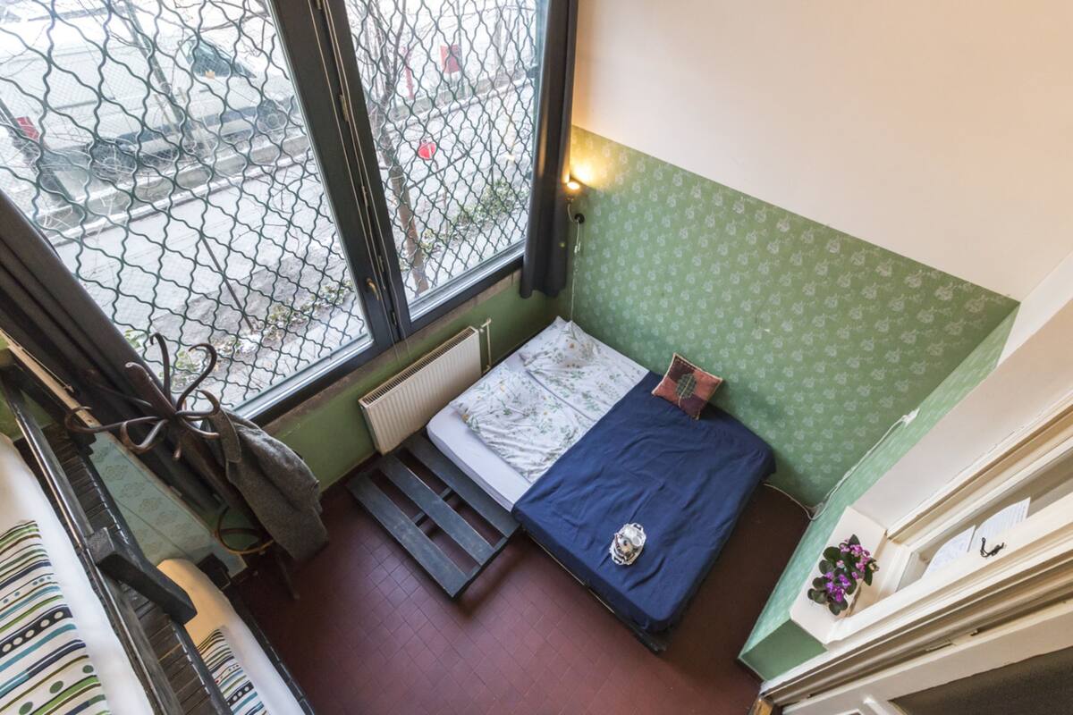 A small, compact room with a bed, a radiator, a window with a view of the street, and a green patterned wall.