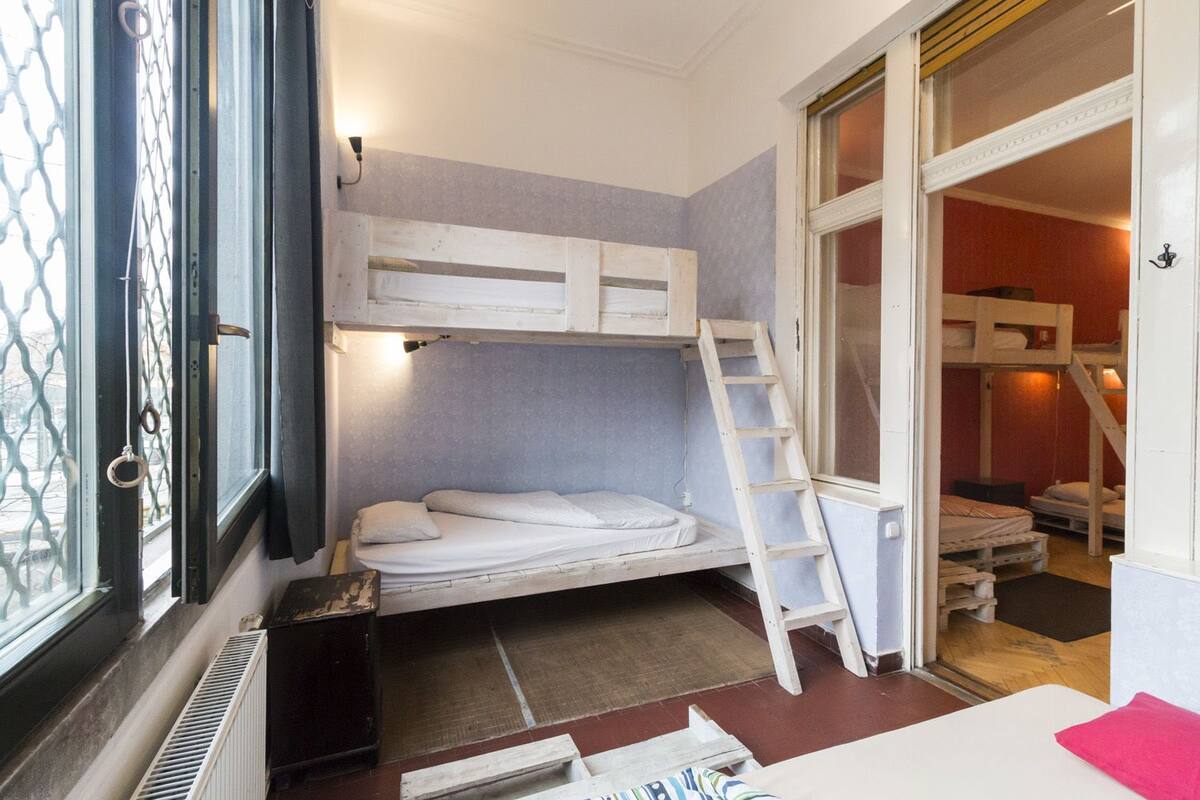 A bunk bed room with a ladder, a window with a metal grill, and a door leading to another room.