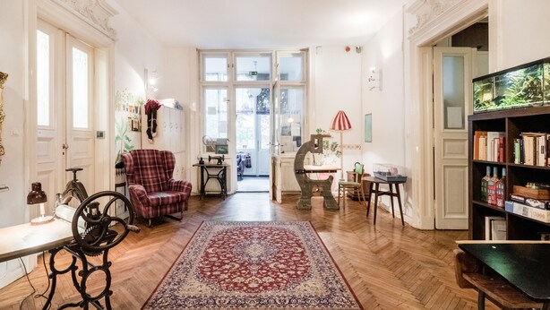 Een ruime woonkamer met een groot raam, een rood tapijt met patroon, een geruit fauteuil, een vintage naaimachine en een boekenkast.