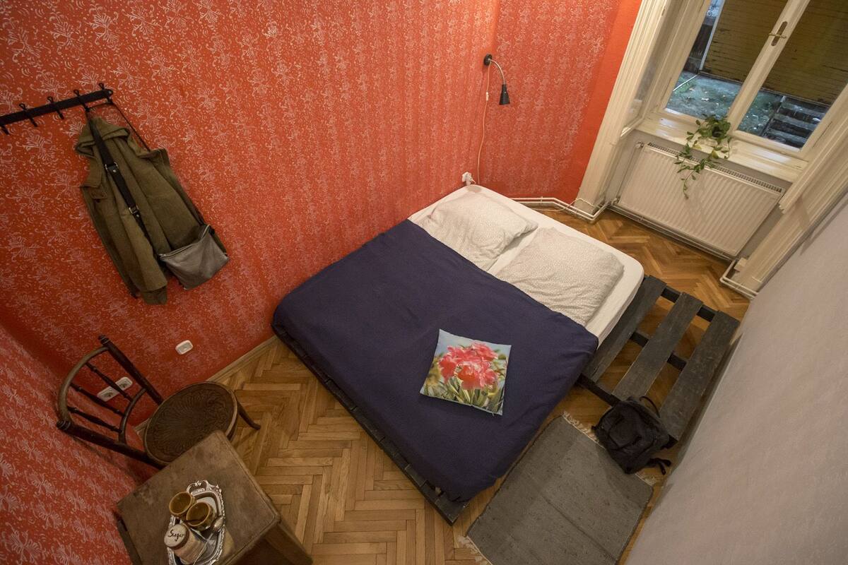 A small bedroom with a bed, a chair, a small table with cups, and a window with a view.
