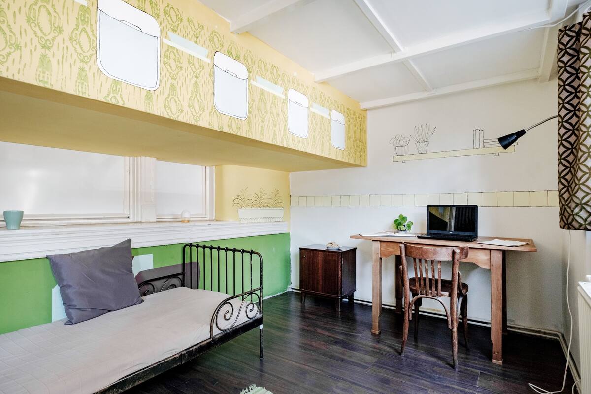 A compact room with a bed, a desk with a laptop, a small cabinet, and a window with a view of plants.