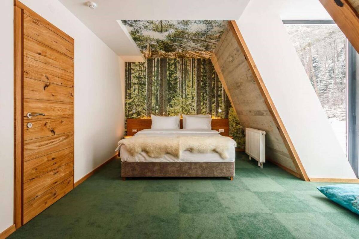 A bedroom with a sloped ceiling, a bed with a white blanket, a wooden wardrobe, and a forest-themed wall.