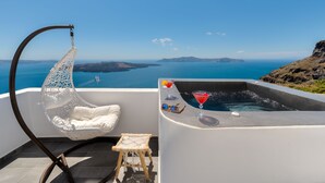 A balcony with a white hanging chair, a small stool, and a hot tub with drinks and sunglasses. - Katris Apartments (Santorini)