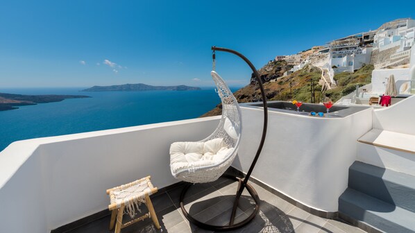 Une chaise en osier blanche sur un balcon, avec vue sur la mer et sur un bâtiment blanc doté d’un escalier.